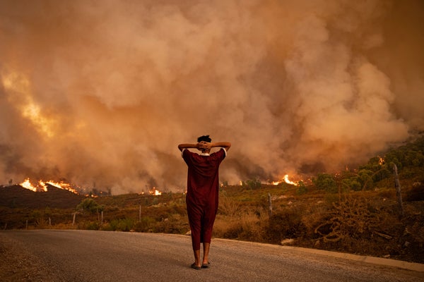 Backview of a woman in a dark red long dress and slippers crossing arms behind head looking a a wildfire landscape.